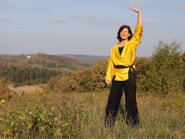 Man sieht eine Frau bei einer Übung in der Natur: Eine Hand oben, die andere unten. Sie lächelt sanft. Ihr Blick geht in die Weite.